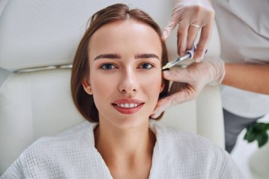 Mutlu zarif kadın ilaç sandalyesinde uzanıyor ve güzellik kliniğinde hiyalüronik asit cilt bakımı oluyor.