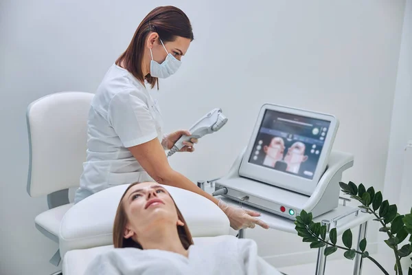 Cosmetólogo morena en uniforme blanco mirando a la pantalla del dispositivo moderno en clínica de belleza — Foto de Stock