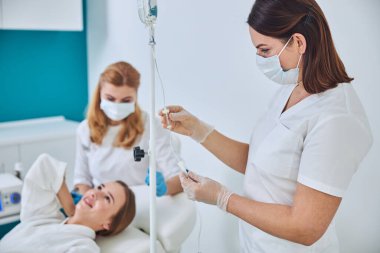 Brunette physician in white uniform and gloves looking to the intravenous vitamin drip in spa salon