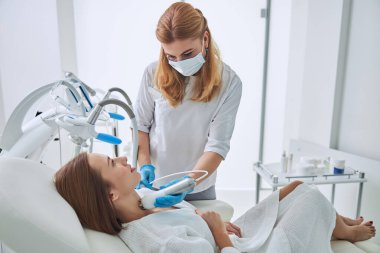 Young pretty lady in white bathrobe sitting in medicine armchair and looking to the beautician during the beauty treatment in spa salon