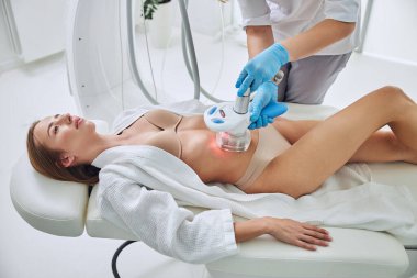 Charming adorable female lying on medicine table while getting cavitation body contouring treatment in medicine beauty clinic