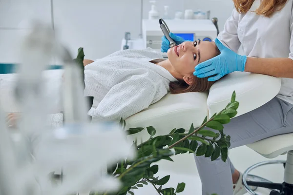 Charming funny Caucasian female lying on medicine chair while receiving rejuvenation facial procedure in beauty clinic — Stock Photo, Image