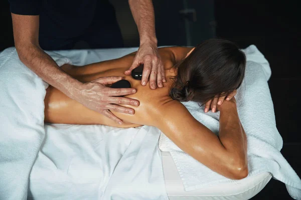 Γαλήνια γοητευτική γυναίκα ξαπλωμένη στο τραπέζι μασάζ, ενώ λαμβάνει επαγγελματική χειροκίνητη διαδικασία στο κέντρο υγείας ευεξίας — Φωτογραφία Αρχείου