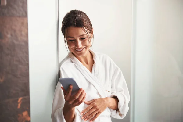 Hermosa mujer caucásica elegante en albornoz de felpa blanca mirando a la pantalla del teléfono celular en el salón de belleza — Foto de Stock