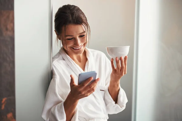 Γέλιο γοητευτική κυρία με κούπα στο χέρι κοιτάζοντας στην οθόνη του κινητού τηλεφώνου στο σπα σαλόνι — Φωτογραφία Αρχείου