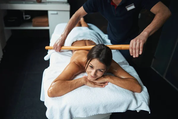 Felice donna bruna allegra sdraiata sul lettino da massaggio mentre ottiene la terapia di massaggio manuale con lana di legno nel salone di bellezza — Foto Stock