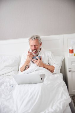 Adult Caucasian man spending time in bedroom at the laptop while eating oatmeal