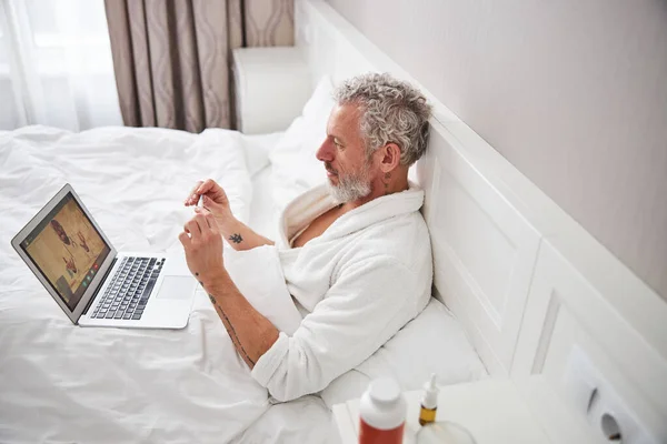 Hombre caucásico adulto en albornoz blanco hablando con el médico en línea mientras está sentado en la habitación interior moderna —  Fotos de Stock