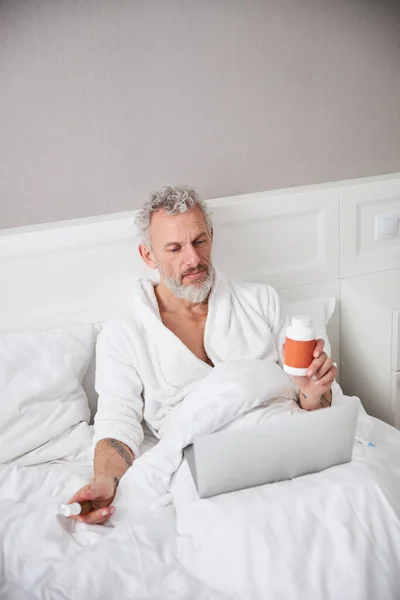 Мужчина в белом халате держит банку с оранжевыми таблетками в руке, сидя на спальне — стоковое фото