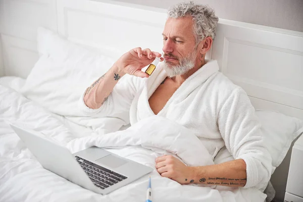 Varón barbudo sentado en la cama mientras gotea la nariz con spray medicinal en la habitación en el interior — Foto de Stock