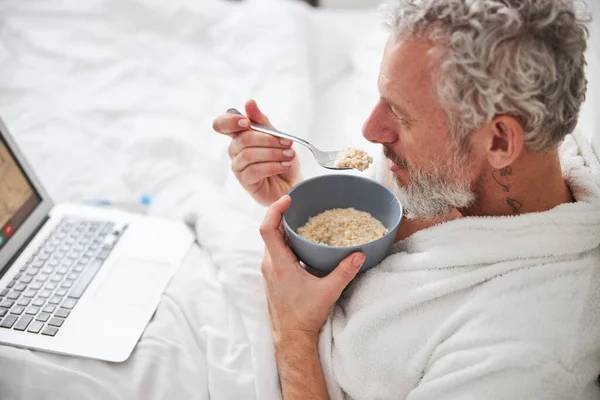 Багатий кавказький самець у білій халаті дивиться на екран ноутбука, коли їсть вівсяну їжу. — стокове фото