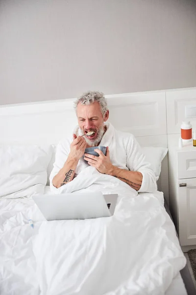 Un bărbat caucazian adult petrece timp în dormitor la laptop în timp ce mănâncă fulgi de ovăz — Fotografie, imagine de stoc