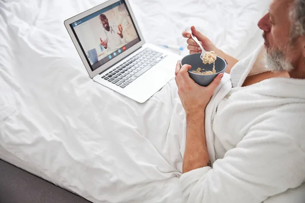 Przystojny brodaty mężczyzna siedzi na łóżku podczas śniadania w mieszkaniu — Zdjęcie stockowe