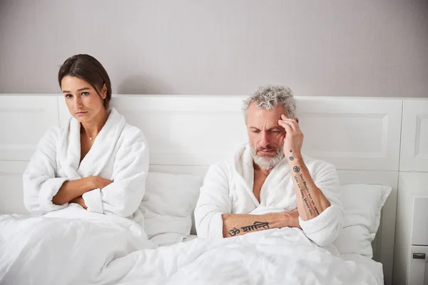 Exited brunette woman and thoughtful bearded man in white bathrobes looking away in bedroom — ストック写真