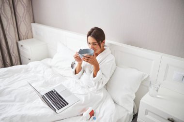 Charming young lady spending time on bed at the laptop and tasty meal in home apartment