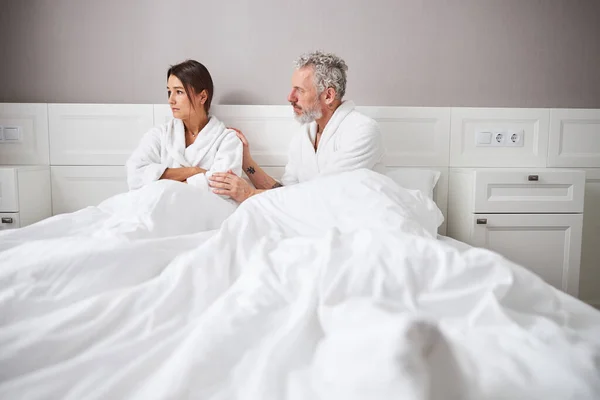 Sad stressed woman in white bathrobe looking away while sitting on the bed in home apartment — Stock fotografie