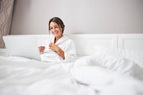 Joven bonita dama en albornoz de felpa blanca utilizando un dispositivo moderno en el dormitorio — Foto de Stock