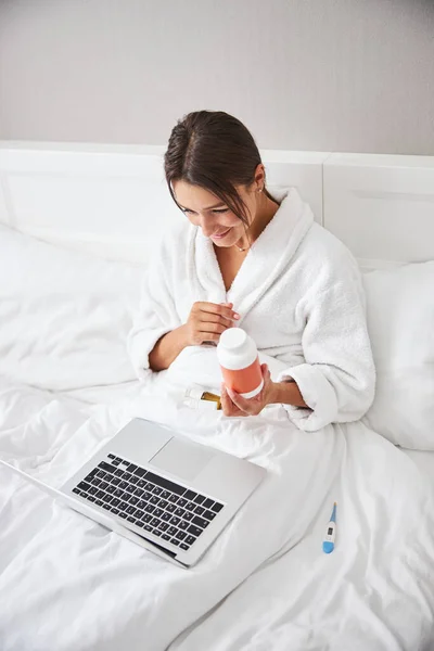 Encantadora mulher caucasiana elegante falando on-line na internet enquanto segurando frasco pílula nas mãos — Fotografia de Stock