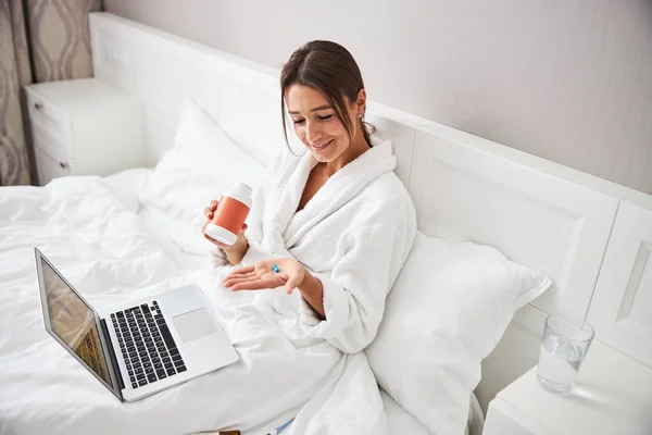 Atractiva mujer caucásica sentada en la cama en el portátil y mirando a la píldora en el brazo —  Fotos de Stock