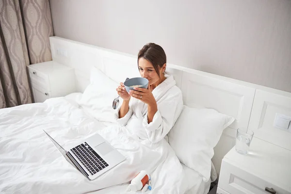 Encantadora joven pasar tiempo en la cama en el ordenador portátil y sabrosa comida en el apartamento en casa — Foto de Stock