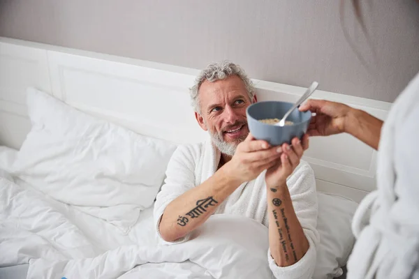 Hombre adulto guapo mirando hacia otro lado mientras sostiene el tazón con cereal en el hogar habitación interior moderna — Foto de Stock