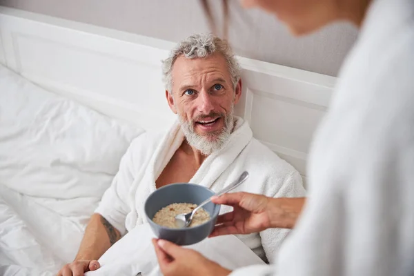 Barbă caucazian de sex masculin petrecerea timpului pe pat în timp ce femeia dând farfurie cu masa în apartament acasă — Fotografie, imagine de stoc