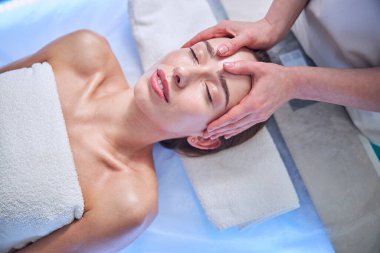 Beautiful elegant female with towel on her body relaxing at the spa face massage in spa salon