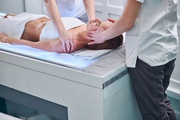 Beautiful Caucasian woman with towel on her body lying on massage table and receiving hand massage on face and shoulder in beauty center — Stock Photo, Image