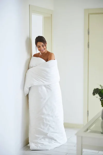 Herrliche junge, lachende Frau in Bettdecke steht im gemütlichen Flur — Stockfoto