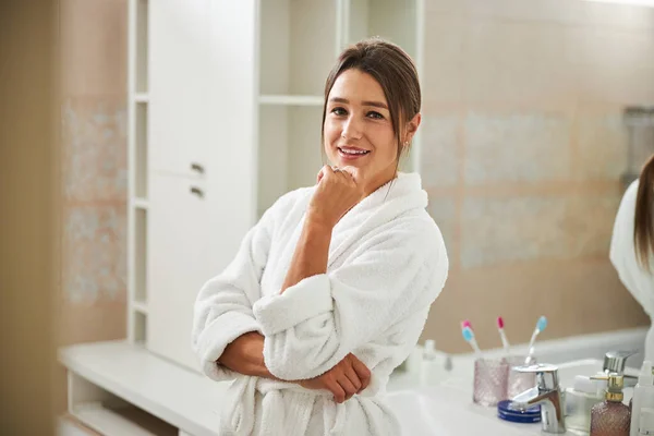 Charmante elegante Frau im Plüschmantel, die im Badezimmer die Hand unter dem Kinn hält — Stockfoto