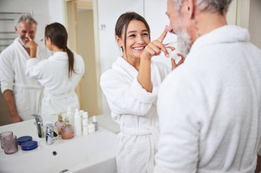 Brunette Caucasian charming female in white bathrobe taking care to her husband in room indoors