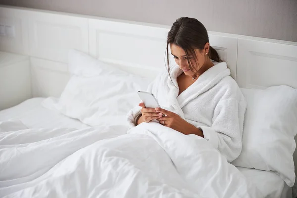 흰색 목욕 복을 입은 아름다운 백인 여성 이 핸드폰을 사용하면서 침대에 누워 있다 — 스톡 사진