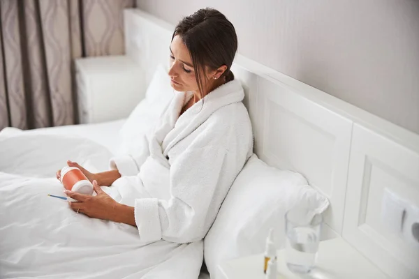 Morena mujer caucásica sentada en la cama con termómetro infrarrojo en las manos en el dormitorio —  Fotos de Stock