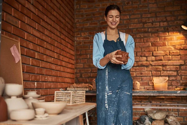 Великолепная молодая ремесленница с глиняной посудой в руках наслаждается процессом работы в керамической мастерской — стоковое фото
