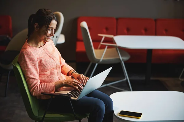 Joven mujer caucásica sonriente en chaqueta rosa charlando en línea mientras navega por Internet en el espacio de trabajo conjunto —  Fotos de Stock