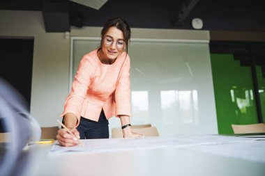 Young Caucasian business lady with pen in hand working with blueprints in the modern cozy office