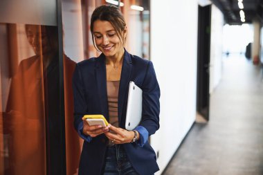 Charming business lady in formal wear chatting on cell phone while holding laptop in hand