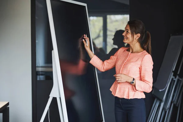Happy attractive caucasian lady in pink jacket standing near the blackboard while writing something — Stockfoto