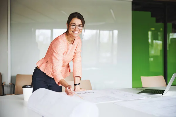 Young attractive Caucasian female business analyst standing near the table and holding pen in hand — Stock Fotó