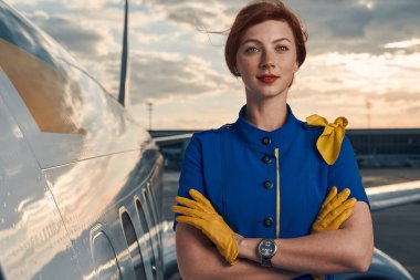 Calm red-haired stewardess posing for the camera by an aircraft clipart