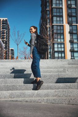 Sırt çantası ve cep telefonu olan neşeli bir turist merdivenlerden yukarı çıkıyor.