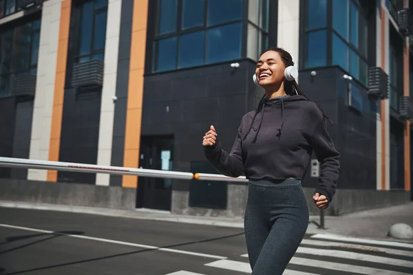 Leende glad idrottskvinna i trådlösa hörlurar och aktiva kläder joggar framåt — Stockfoto