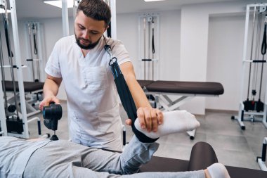 Pediatric physiotherapist with massaging percussion hammer treats girls leg and back with espander