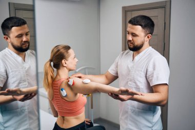 Physiotherapist puts electric stimulator on young woman arm at mirror in modern therapeutic office