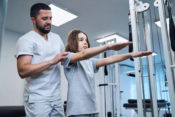 Pediatric therapist checks posture of girl with her arms stretched forward in rehabilitation center — Stock Photo, Image