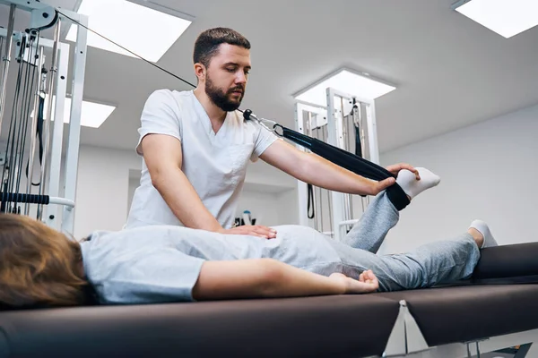 Volwassen fysiotherapeut behandelt knie van jonge vrouwelijke patiënt. Meisje met elastisch verband op voet — Stockfoto