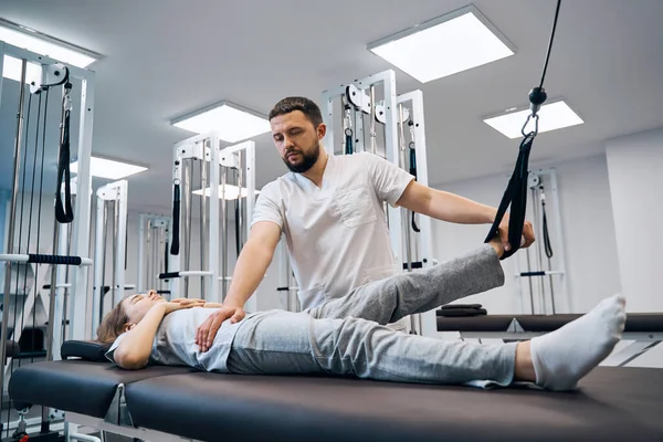 Girl lying on physical therapy bed, doing exersises on elastic strength machines with chiropractor — 스톡 사진