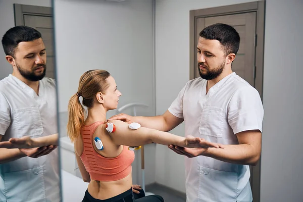 Physiotherapist puts electric stimulator on young woman arm at mirror in modern therapeutic office — стоковое фото