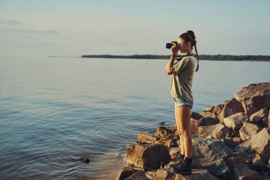 Nehir kıyısındaki sıska kadın su yüzeyinin fotoğraflarını çekiyor.