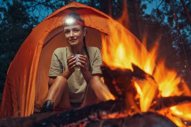Kamp yapan kadın ateşe bakarken içki içiyor.